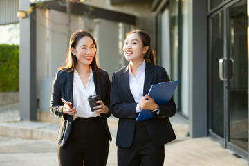 Obraz premium Two smiling Asian businesswomen walking and talking outside a modern office building, representing teamwork and active partnership.