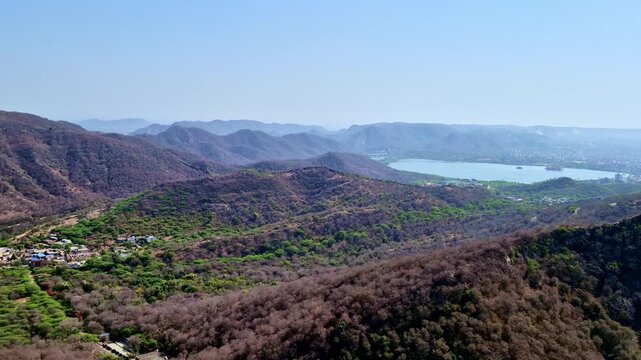 A bright daytime  showing a panoramic view of the Aravalli hills with dry forest valleys, scattered villages, and Maota Lake visible in the distance under a clear blue sky.