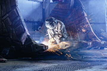 Welder kneeling cutting large excavator bucket inside factory bay, recovery work of industry work © Parilov