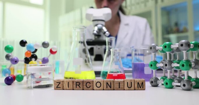 Wooden blocks spell word Zirconium near flasks and molecular structure models. Lab scientist calibrates tools studying properties for industry