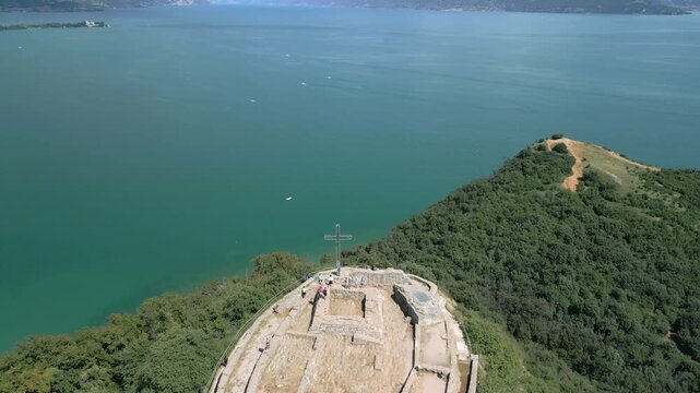 Drone pullback tilt down over Rocca di Manerba del Garda, sunny summer view of medieval fortress ruins and tourists on lookout above Lake Garda turquoise water