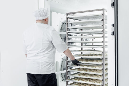 Male pastry chef in confectionery industry places puff pastry buns in freezer for shock freezing