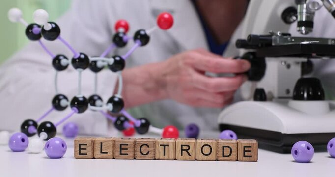 Wooden blocks spell word Electrode near microscope and molecule model. Laboratory worker adjusts equipment for electrical testing and analysis