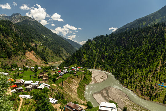 View of a village nestled between towering, verdant mountains, bisected by a milky river under a bright sky, Kel, Azad Kashmir, Pakistan.