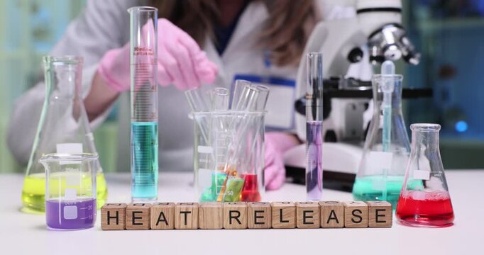 Wooden blocks spell phrase Heat Release at flasks and test tubes. Scientist mixes colorful liquids measuring temperature during reaction by microscope