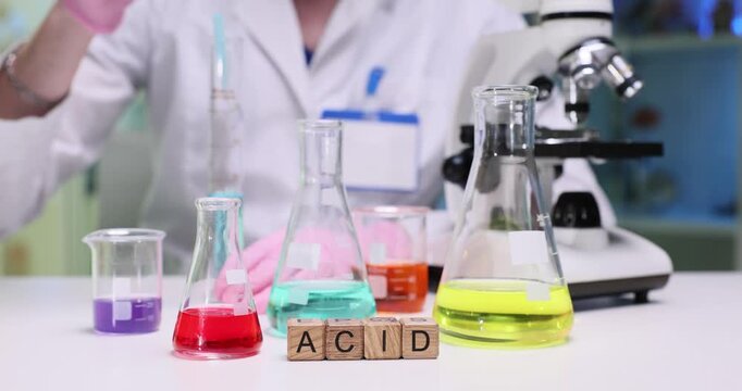 Wooden blocks spell word Acid near flasks with colorful liquids. Scientist mixes solutions measuring reaction while lab glassware stays sterile