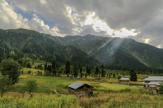 View of rustic wooden houses nestled in a vibrant green meadow against a backdrop of majestic mountains and a cloudy sky, Arang Kel, Azad Kashmir, Pakistan.