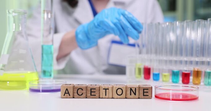 Wooden blocks spell word Acetone near test tubes in rack and lab tools. Gloved scientist handles samples recording data during chemical analysis