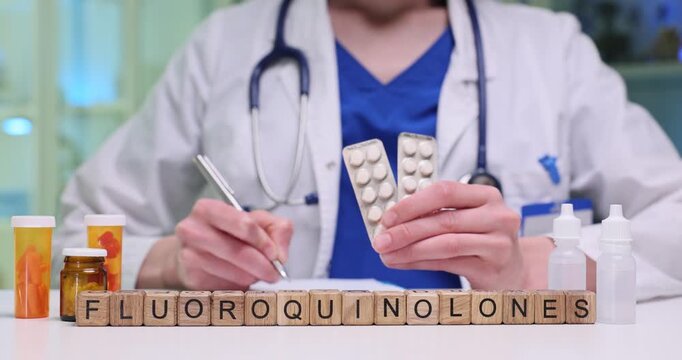 Wooden blocks spell word Fluoroquinolones near pills and stethoscope on desk. Doctor holds blister packs writing dose notes for patient treatment