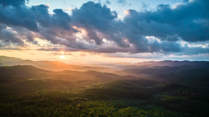 Obraz premium Aerial view breathtaking sunset over forest. Sun's rays break through dramatic clouds, casting golden glow across rolling hills and valleys. Distant mountains creating serene and picturesque scene.