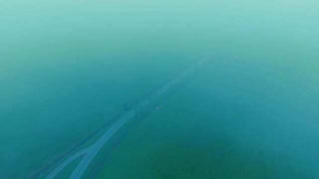 Winding rural road appearing through dense fog above countryside pasture. Low clouds drifting across agricultural fields while aerial perspective revealing mist covered farmland. Moody landscape with