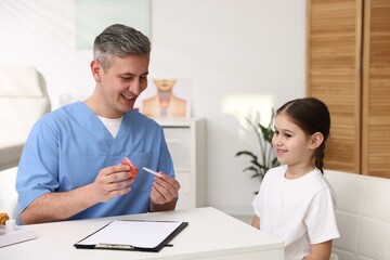 Obraz premium Smiling endocrinologist showing model of thyroid gland to his little patient at white desk in clinic