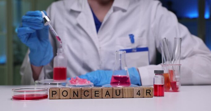 Wooden blocks spell word Ponceau 4R near dye vial and flask. Lab scientist pipettes colorant dissolving powder while measuring dose for additive test
