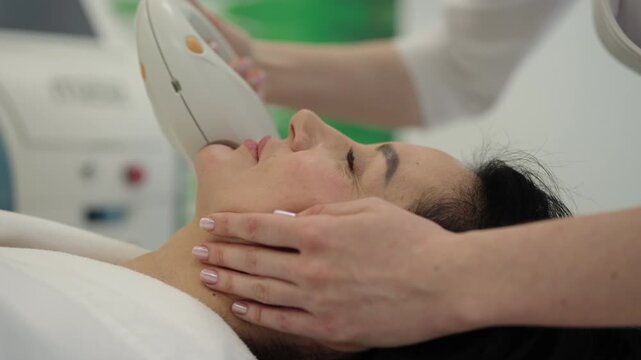 Closeup of middle-aged woman receiving relaxing rf lifting procedure for face and neck in beauty salon, cosmetologist using professional equipment for non invasive facial treatment.