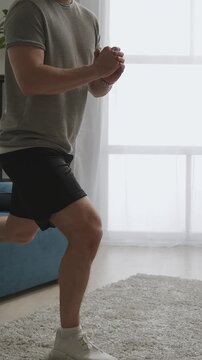 Focused man performing single-leg Bulgarian split squat exercise at home in bright living room, building lower body strength, balance, and core stability without equipment