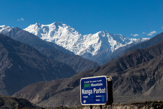 View of the majestic Nanga Parbat sign surrounded by rugged, dark mountains against a bright blue sky, snow-capped peaks piercing above, Astore, Gilgit, Pakistan.