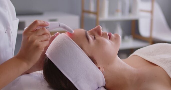 Female client facial treatment beautician hands applying anti age device, dermapen microneedling pen on forehead, eyes closed in comfort, professional care rejuvenation, lying relaxed on treatment bed