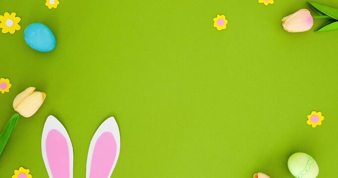 White bunny ears, colorful Easter eggs, tulips and small flowers on a green background with space for text. Spring composition, top view. Looped 4k stop motion animation
