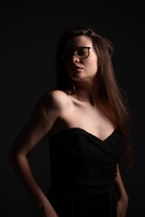 Naklejka premium Young woman with long brown hair wearing fashionable black strapless dress and stylish yellow glasses, posing against a dark background, portraying elegance and mystery with subtle light