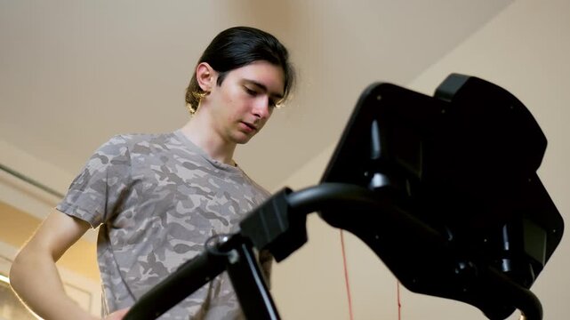 Low angle view of young man training on treadmill during home fitness session in apartment interior. Building endurance through steady cardio workout and focused running pace on indoor exercise