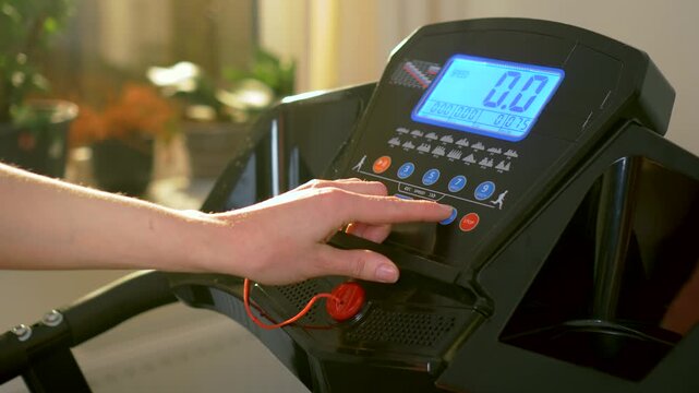 Female hand pressing treadmill control panel while selecting cardio training program at home. Close view adjusting speed and workout settings on digital treadmill console in apartment fitness space