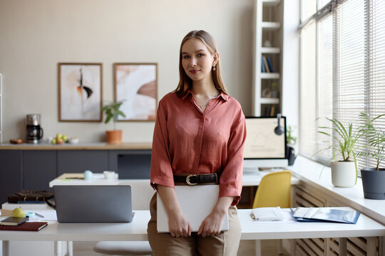 Portrait of young adult businesswoman holding laptop and standing in modern office, presenting professional confidence. Ideal for corporate branding, recruitment, leadership content