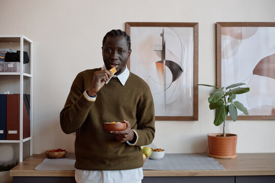 Portrait of Black young adult man eating chips while standing in office kitchen, suggesting casual snacking and everyday lifestyle. Useful for food habits, office life, diet content