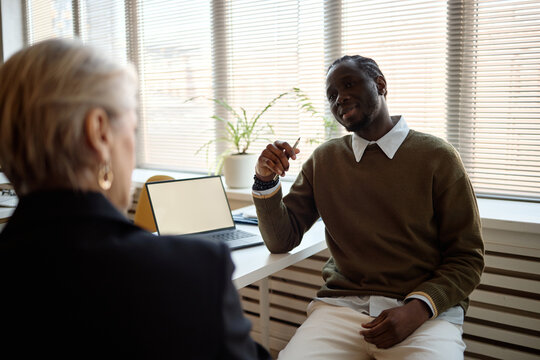 Medium shot of Black young adult man discussing with senior woman across desk, holding pen during office meeting. Supporting business consulting, HR interview, mentoring or coaching content
