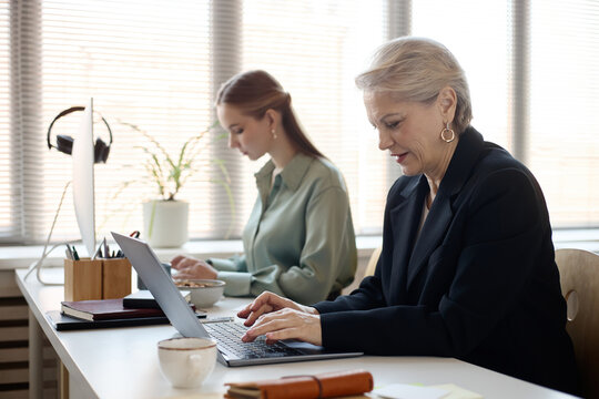 Senior businesswoman working on laptop beside young adult colleague in office, managing tasks and collaboration. Suitable for corporate teamwork, leadership, mentoring, productivity marketing