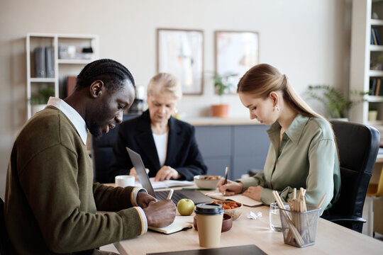 Black young adult man and Caucasian young adult woman taking notes during office meeting while senior colleague working on laptop. Useful for teamwork, planning, corporate training campaigns