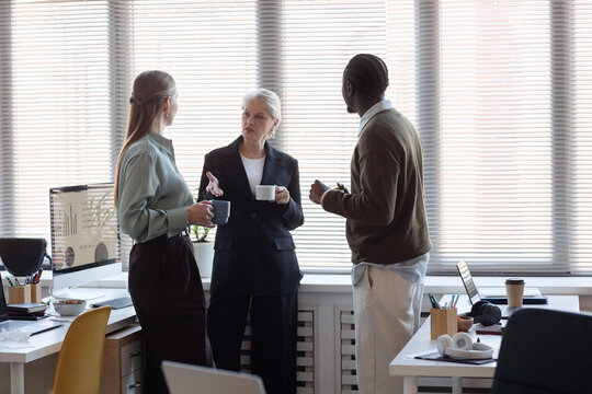Senior businesswoman briefing young female colleague and Black man during office coffee break. Supporting teamwork, leadership communication, workplace culture campaigns