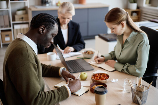 Diverse coworkers taking notes during office meeting, reviewing laptop content and planning tasks. Useful for business teamwork, strategy, productivity, collaboration marketing