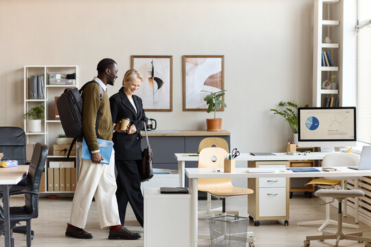 Black young adult man and senior Caucasian businesswoman walking through office, discussing work and carrying laptop folder. Ideal for onboarding, mentorship, teamwork, corporate culture marketing