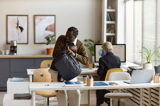 Black young adult man unpacking backpack at office desk while coworker working at computer, suggesting commute or hybrid work. Ideal for workplace mobility, office routine, productivity