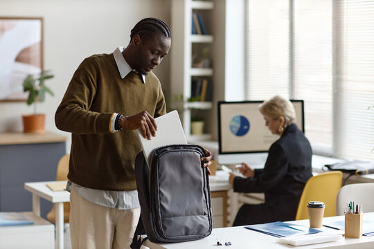 Medium long shot of Black young adult man packing laptop into backpack in office while colleague working at desk. Supporting hybrid work, commuting, business productivity, team workflow