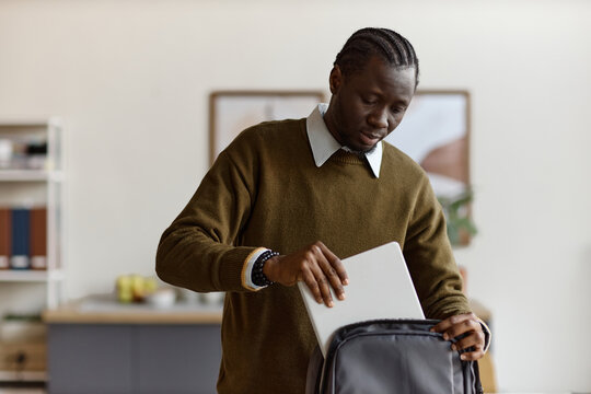Black young adult man packing laptop into backpack, preparing for commute or remote work transition at home. Useful for hybrid work, mobility, productivity, or digital lifestyle marketing