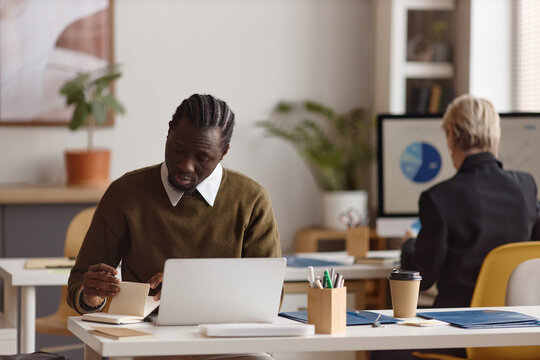 Black young adult man working on laptop and taking notes at office desk while colleague reviewing chart in background. Ideal for business productivity, planning, workplace content