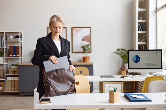 Senior businesswoman packing laptop into bag in office, preparing for workday transition or meeting. Ideal for corporate communication, leadership, productivity themes