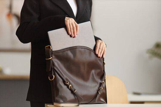 Mid shot of businesswoman packing laptop into shoulder bag, preparing for office commute or meeting. Suitable for corporate mobility, hybrid work, productivity, or career planning ads