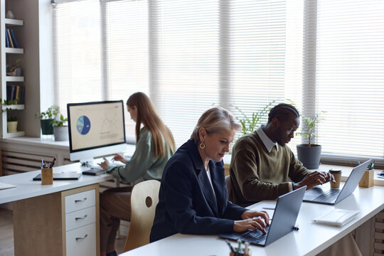 Diverse office team working on laptops and desktop computer, analyzing charts and typing at desks. Suitable for business productivity, teamwork, corporate planning