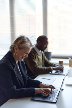 Vertical shot of cheerful senior businesswoman typing on laptop in office while Black male colleague working in background. Supporting corporate teamwork, leadership, training, or productivity marketi