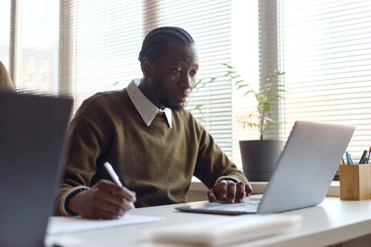 Black young adult businessman working on laptop and taking notes at desk in office, representing focused productivity and planning. Ideal for business, training content