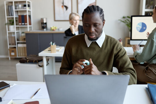 Black young adult businessman working on laptop in office, holding grip ring and managing pressure while colleagues collaborating behind. Useful for workplace mental health or burnout campaigns