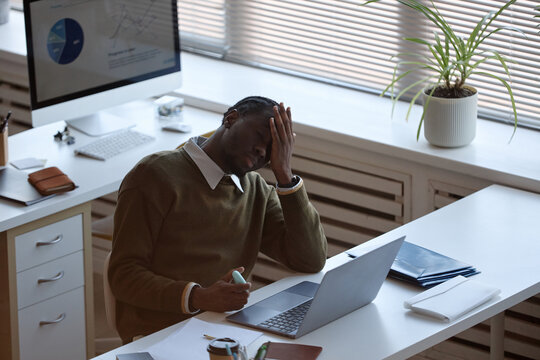 Black young adult businessman holding head while working on laptop at office desk, showing stress and fatigue. Useful for burnout, workload pressure, mental health, corporate wellness themes