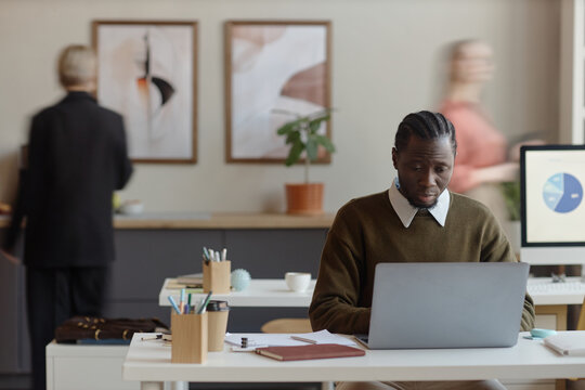 Black young adult man working on laptop at office desk, reviewing business data and multitasking. Suitable for workplace productivity, corporate planning content