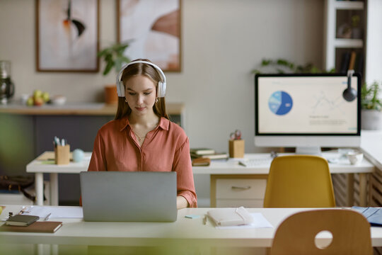 Young adult woman wearing headphones working on laptop at desk, managing online tasks in office. Ideal for office work, productivity, digital business, marketing use