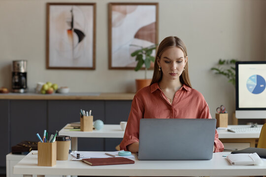 Young adult woman working on laptop at office desk, managing digital tasks and business workflow. Ideal for office work, productivity, corporate communication, startup marketing