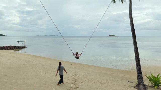 Aerial view of the Bobby Beach shoreline, displaying palm trees, swings, and tranquil turquoise waters meeting the sandy shore, Karimunjawa, Central Java, Indonesia.