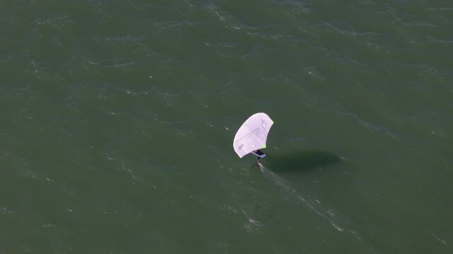 Aerial view of a single wing foiler gliding across the rippled water, a dance of white sail and dark shadow, a serene seascape, Cascais, Lisbon, Portugal.
