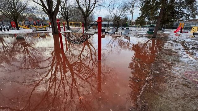 Spring arrives all at once, swallowing a public playground in reddish meltwater until the familiar play space turns into a reflective flooded zone no one can use.